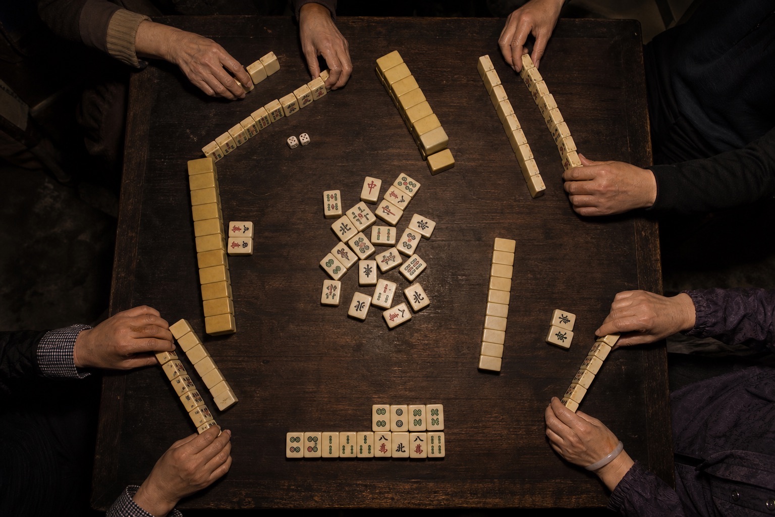 Hands arranging mahjong tiles around a dark wooden table.